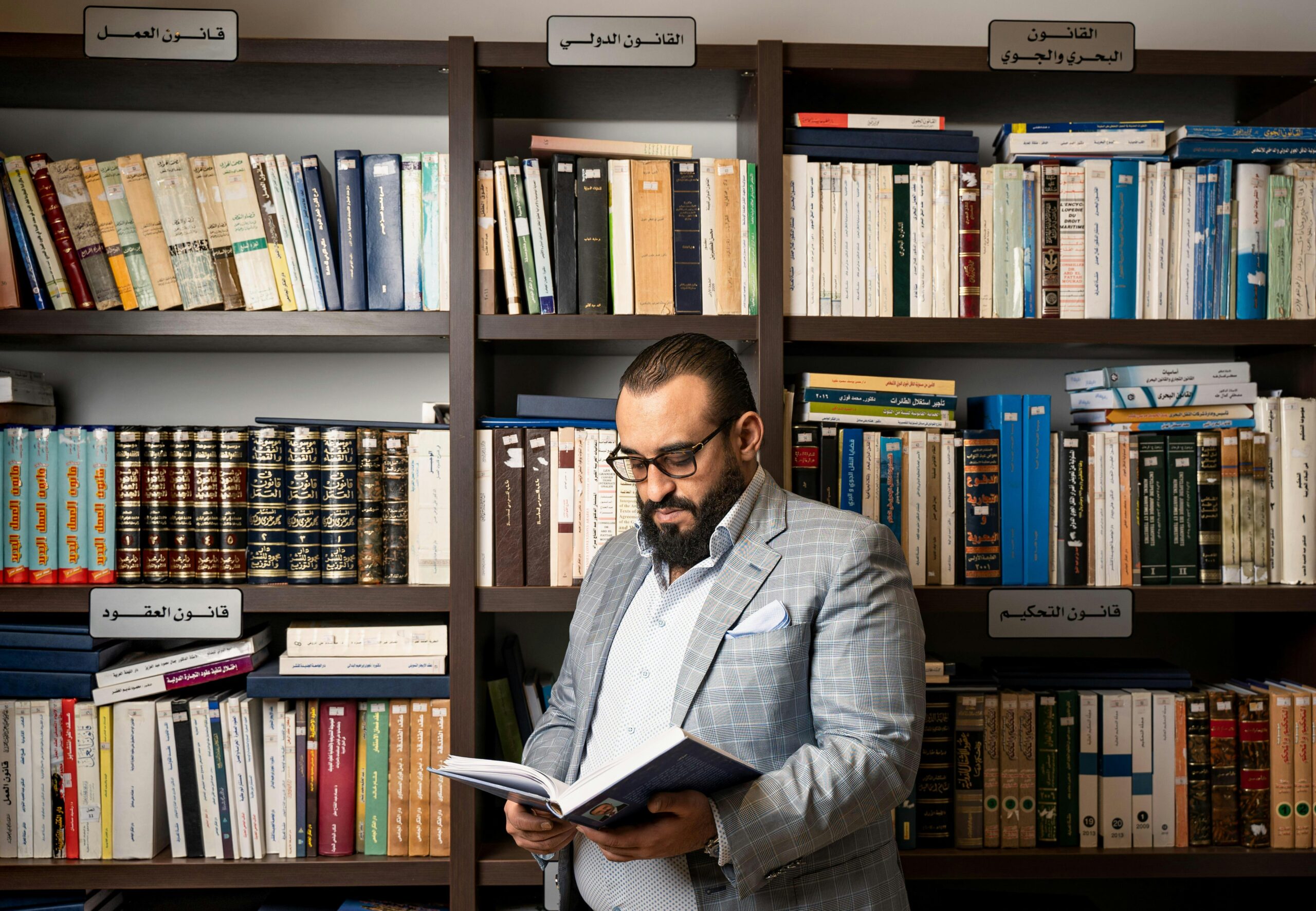 A bearded businessman in a suit reads a book in a library with shelves full of books.